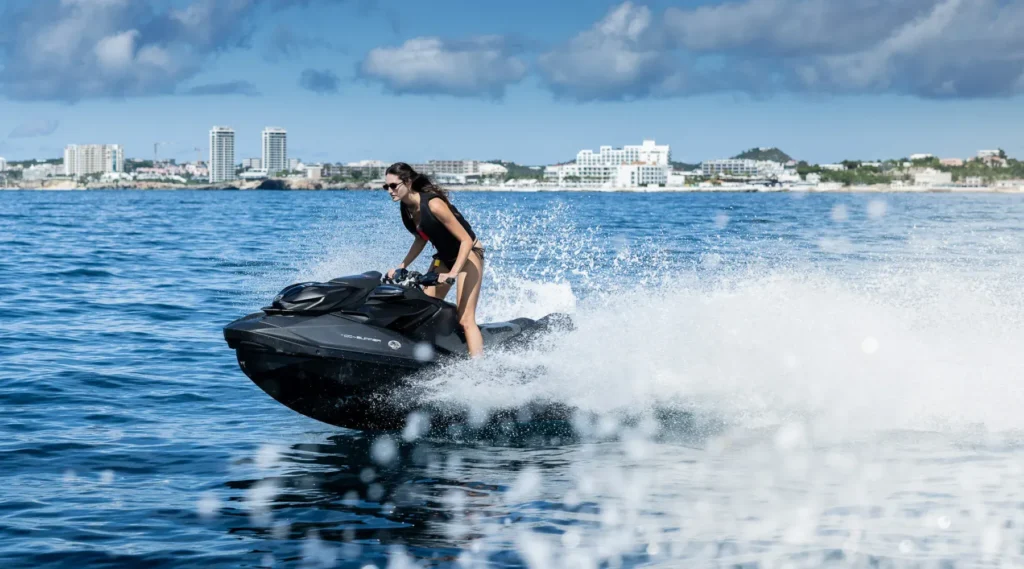 Woman in swimsuit rides black jet ski on blue water near city, with Lurssen CC-Summer yacht for charter in the background.