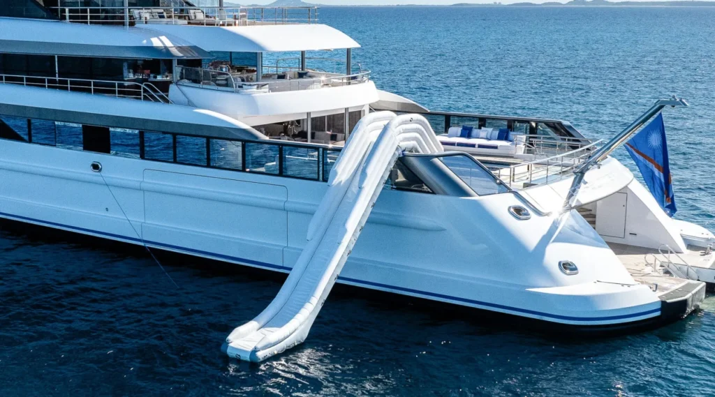 Luxury Lurssen CC-Summer Yacht for Charter with an inflatable slide from the upper deck into the blue ocean, flag at stern.