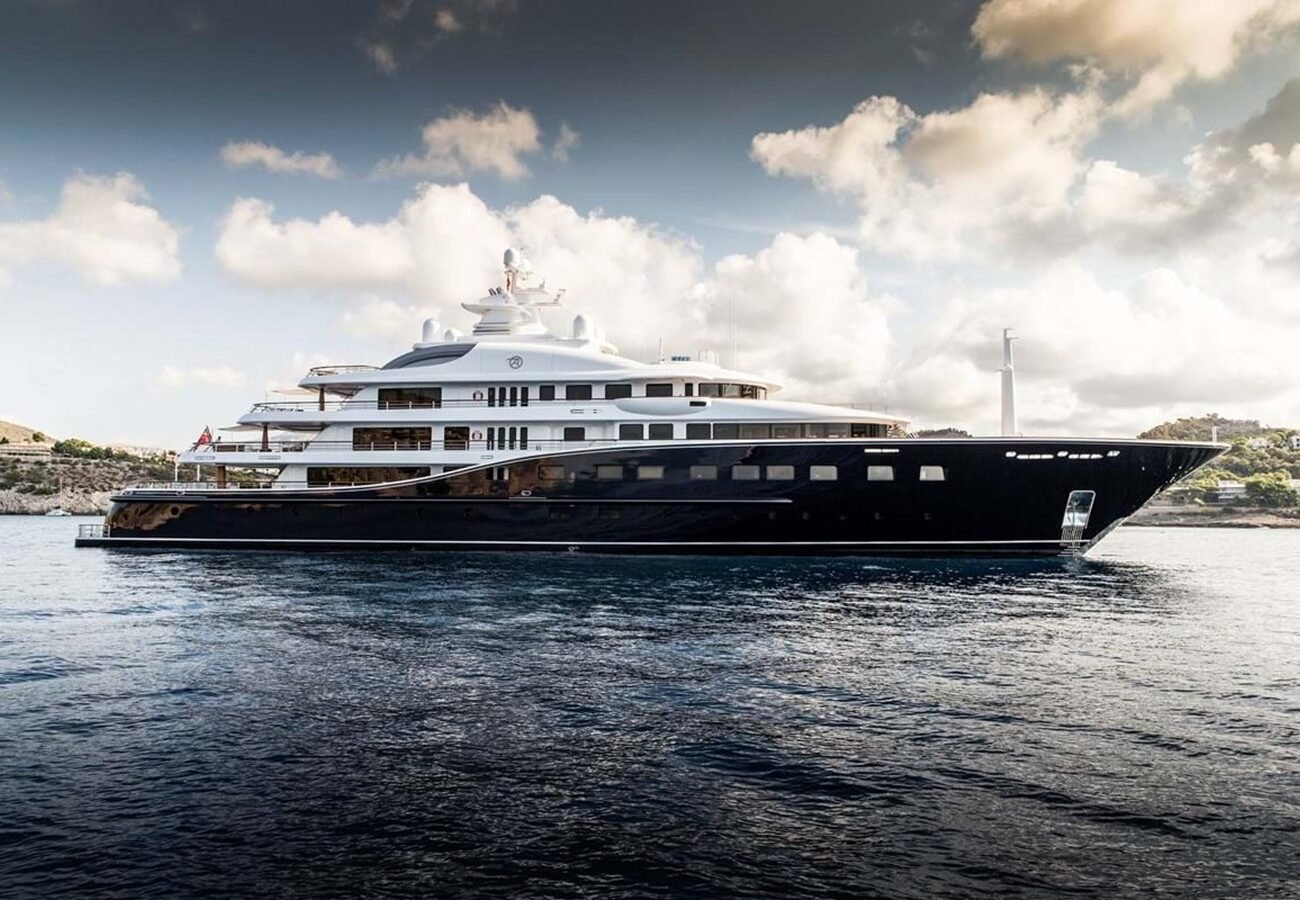 A large, luxurious Derecktor Aquila Yacht for Charter is anchored on calm water under a partly cloudy sky near a rocky coastline.