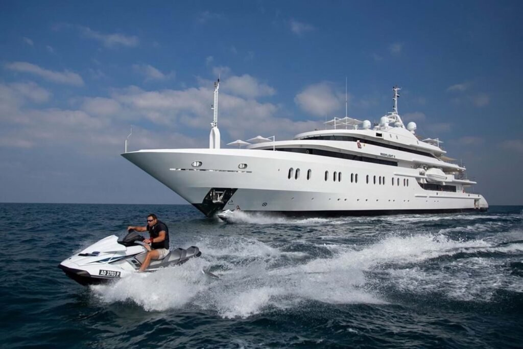 A man rides a jet ski in front of the Neorion Moonlight II Yacht for Charter on calm blue water under a partly cloudy sky.