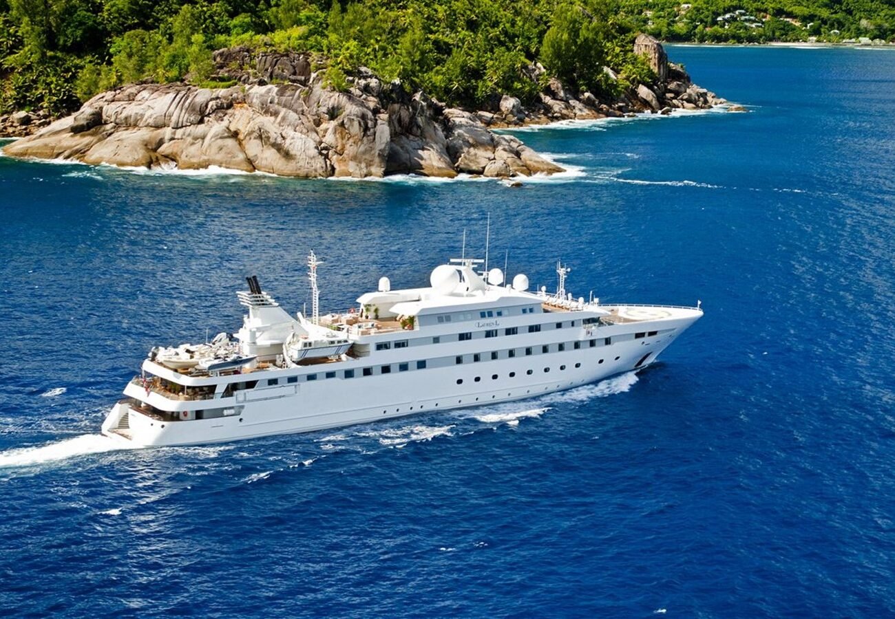 The Cassens-Werft PV Lauren L Yacht sails on deep blue water near a rocky, tree-covered island under a clear sky.
