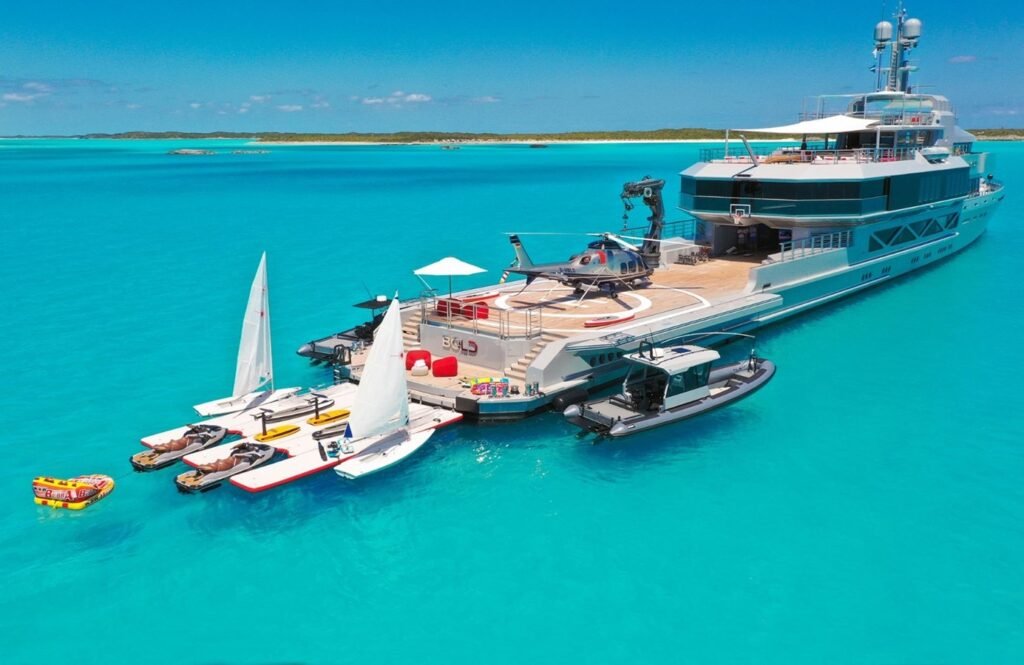 Silver Yachts Bold Yacht for Charter, with sailboats, jet skis, and a helicopter beside its deck in clear blue water.
