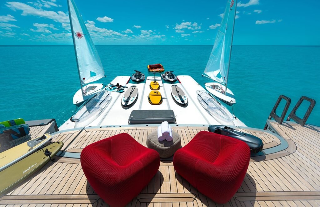 Two red chairs on the Silver Yachts Bold Yacht for Charter, with kayaks, paddleboards, and two sailboats on calm blue water.