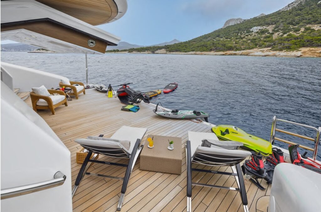 Two lounge chairs face the sea on the Nobiskrug Lady Vera Yacht for Charter, with jet skis and hills in the background.