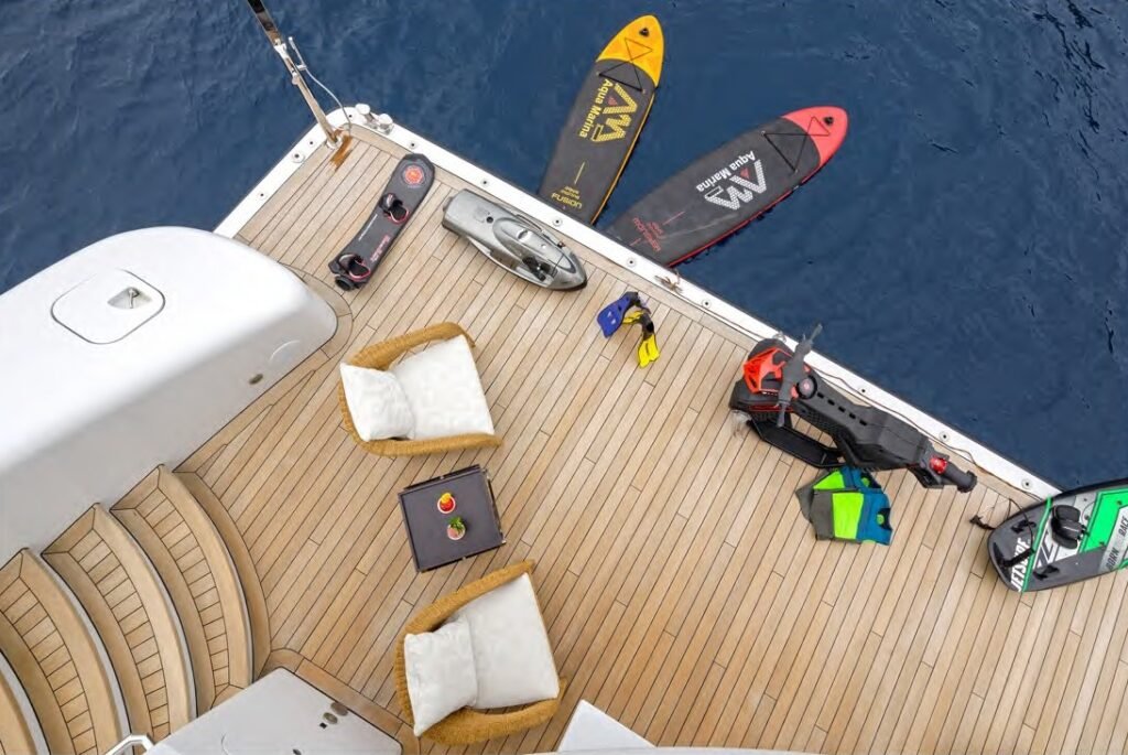 A yacht deck on the Lady Vera with tan chairs, table, steps, water toys, paddleboards, and deep blue water behind.