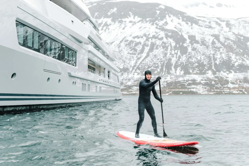 A person in a wetsuit paddleboards near the Abeking Rasmussen Cloudbreak yacht for charter with snowy peaks behind.