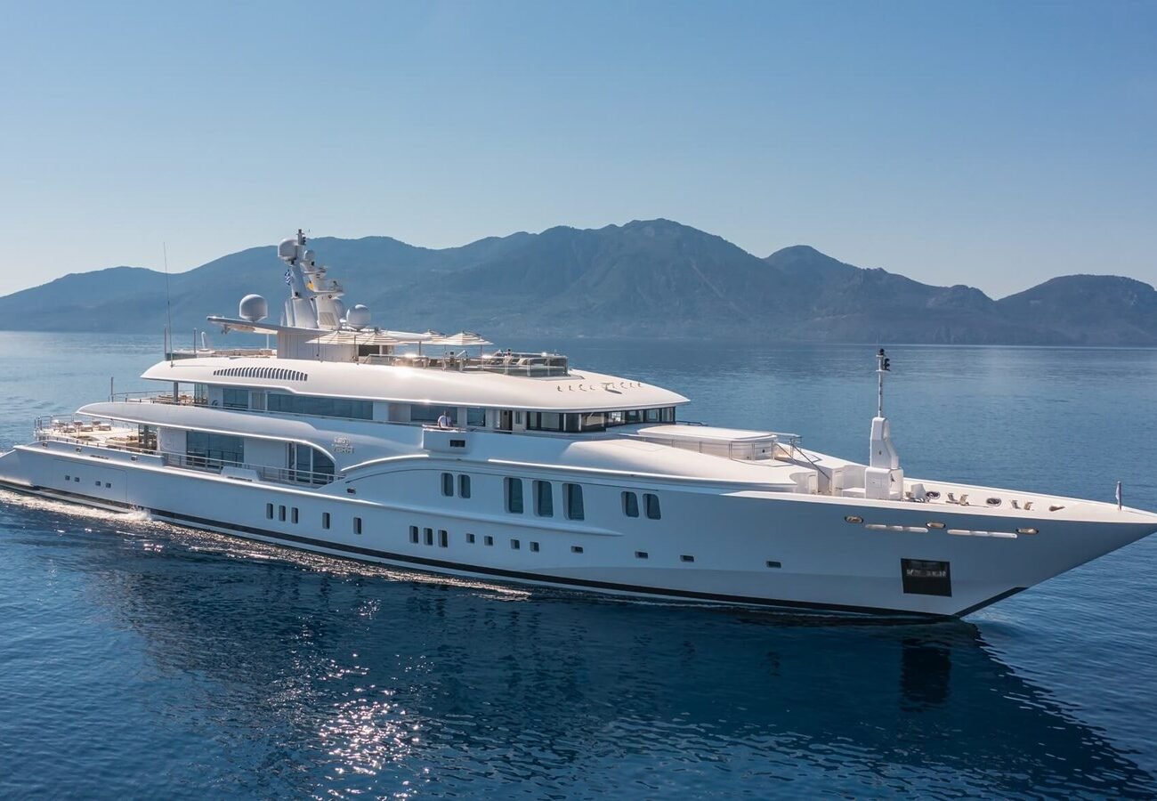 The Nobiskrug Lady Vera Yacht for Charter cruises on calm blue water with mountains in the background under a clear sky.