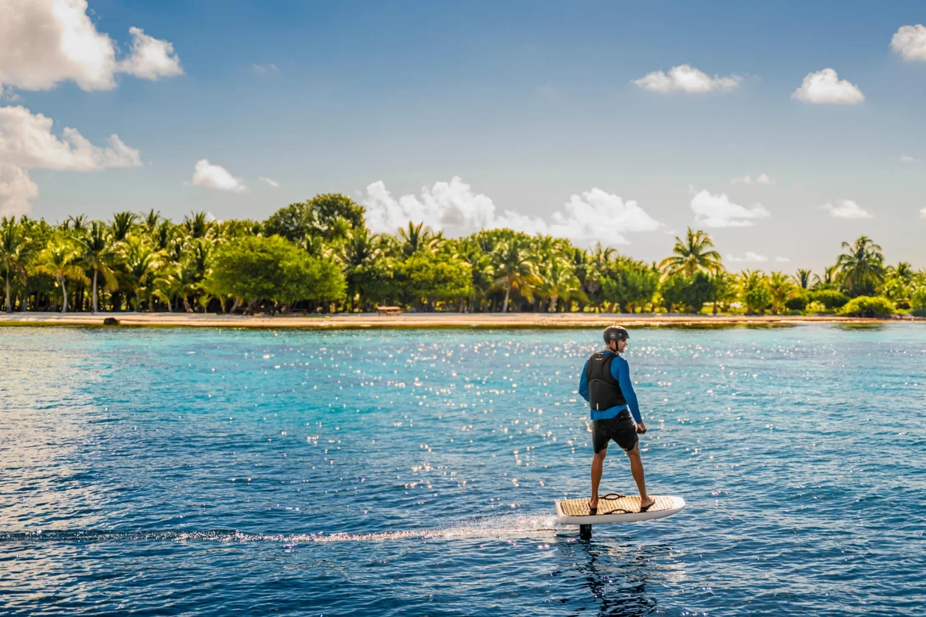 Person rides an electric surfboard on clear blue water near a tropical island with palm trees under a sunny sky.