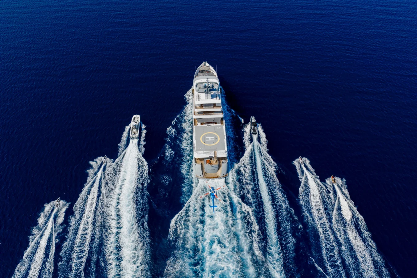 A large yacht with a helipad sails on deep blue water, flanked by three smaller boats creating white wakes.