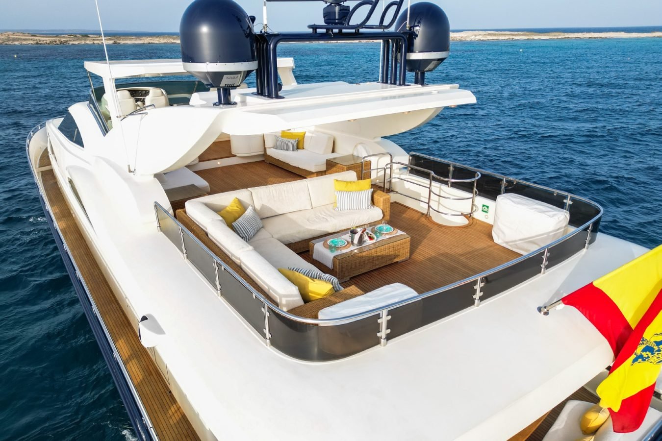 Astondoa 102 GLX My Kirios Yacht For Charter: white yacht, blue water, yellow pillows, wicker table & Spanish flag aft.