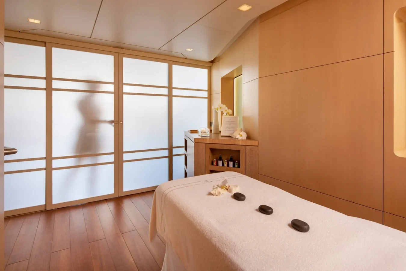 A serene spa room with a massage table, three stones, soft lighting, and a blurred figure behind frosted glass doors.