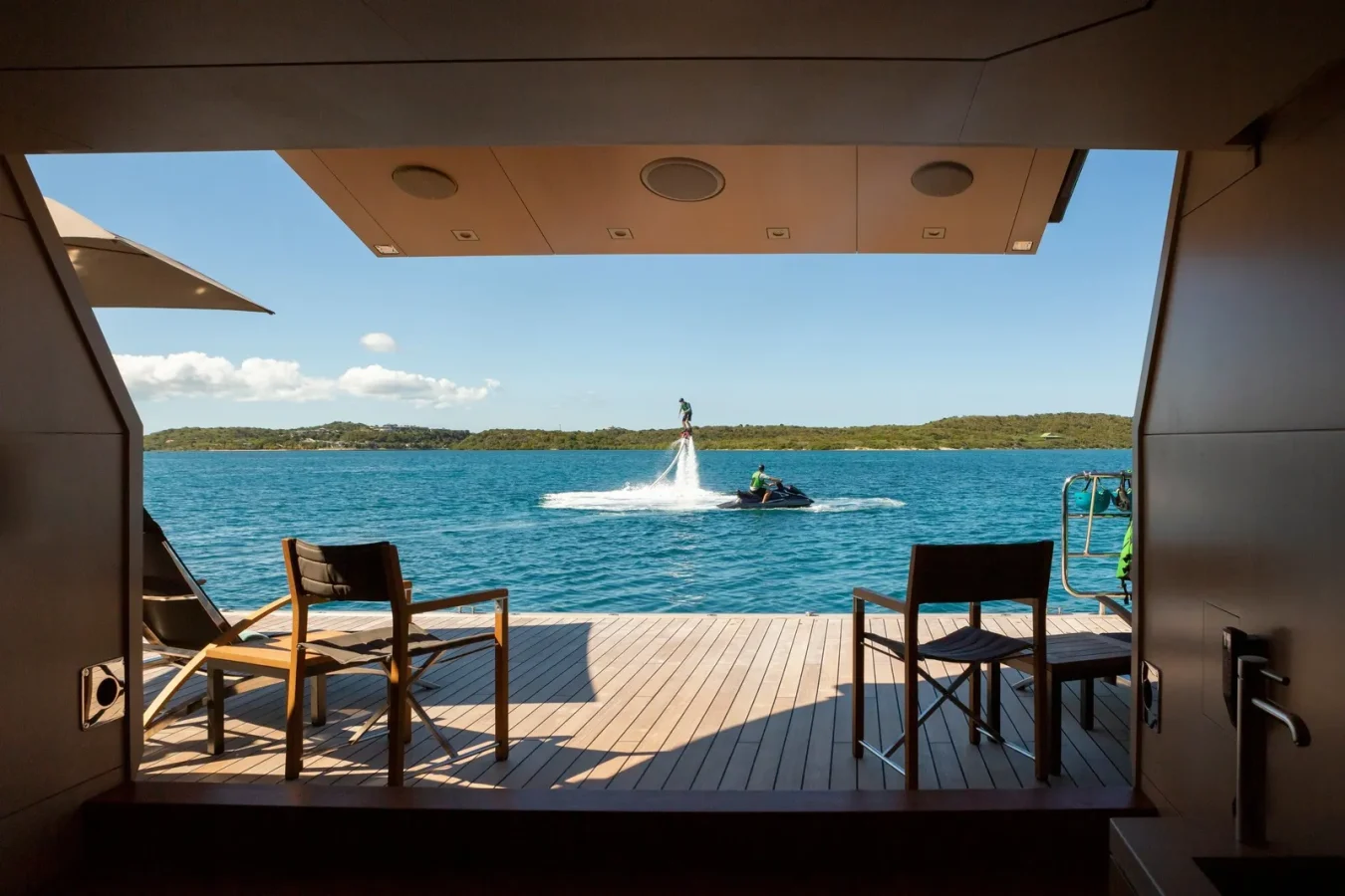 View from a yachts deck: two chairs, calm sea, person on water jet pack, blue sky, green land far away.