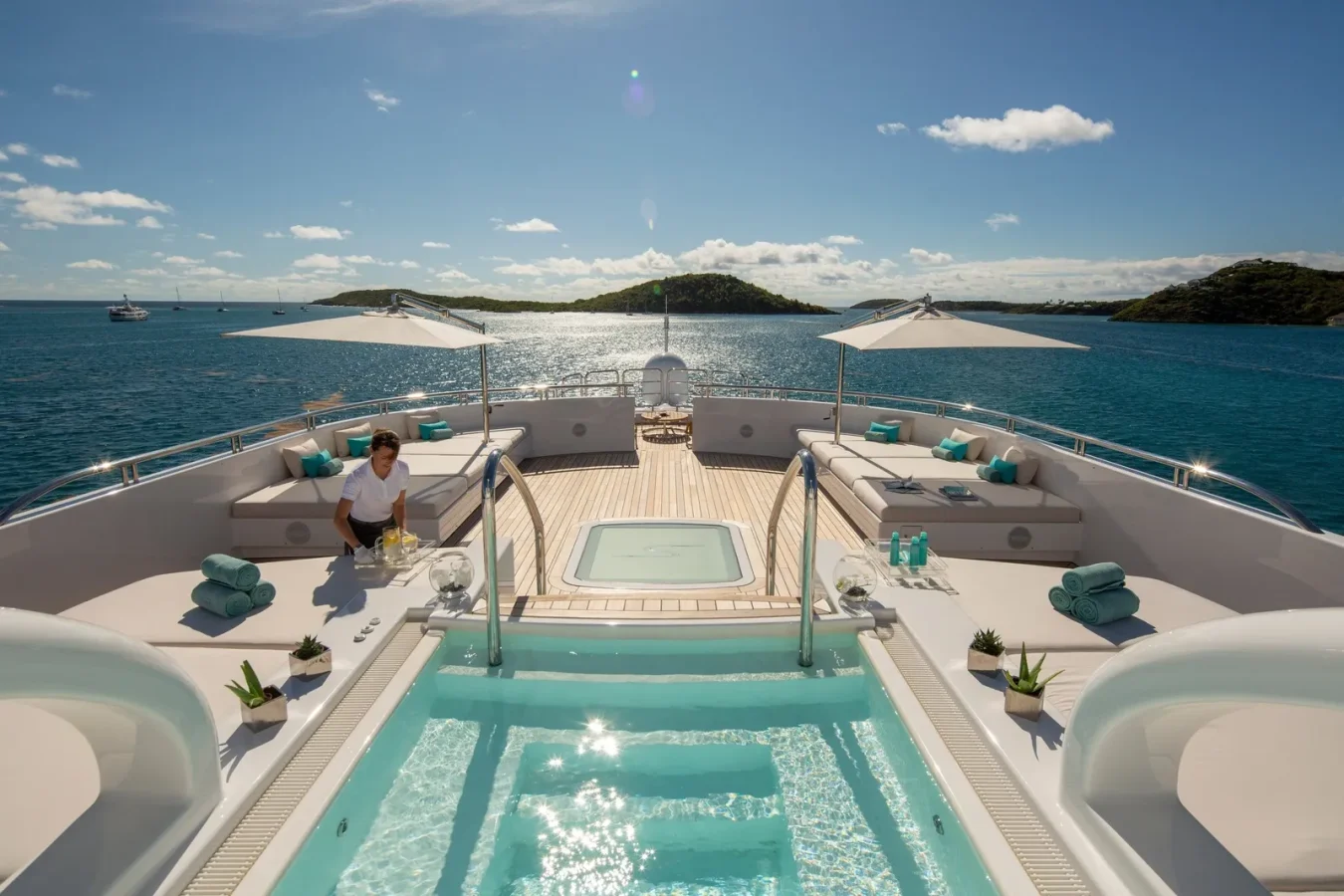 A woman by a yacht pool with blue towels, lounge sofas, umbrellas, and ocean views under a sunny sky.