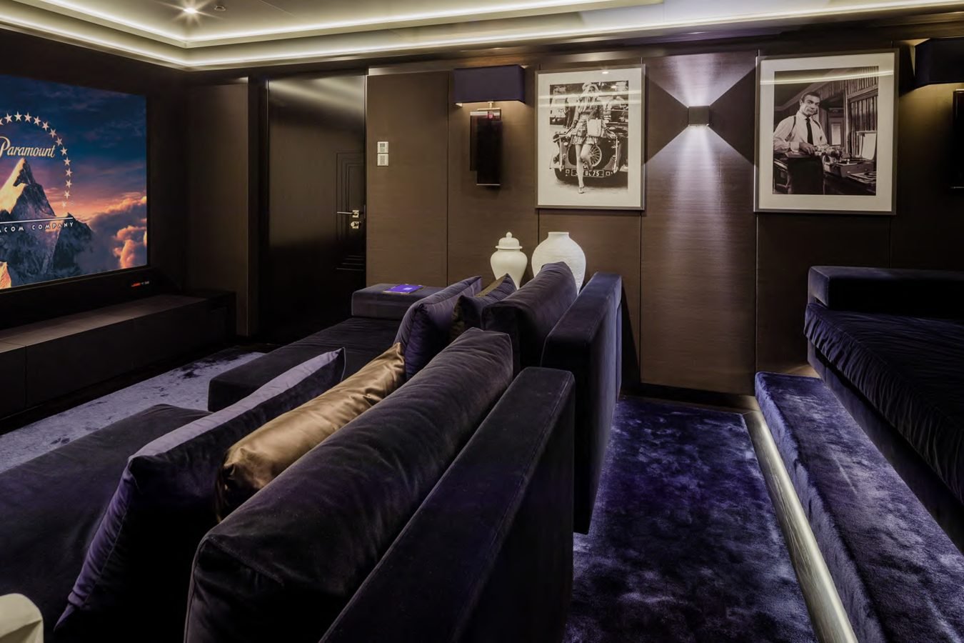 Modern home theater with tiered velvet seats, dark walls, a movie playing, and black-and-white wall photos.