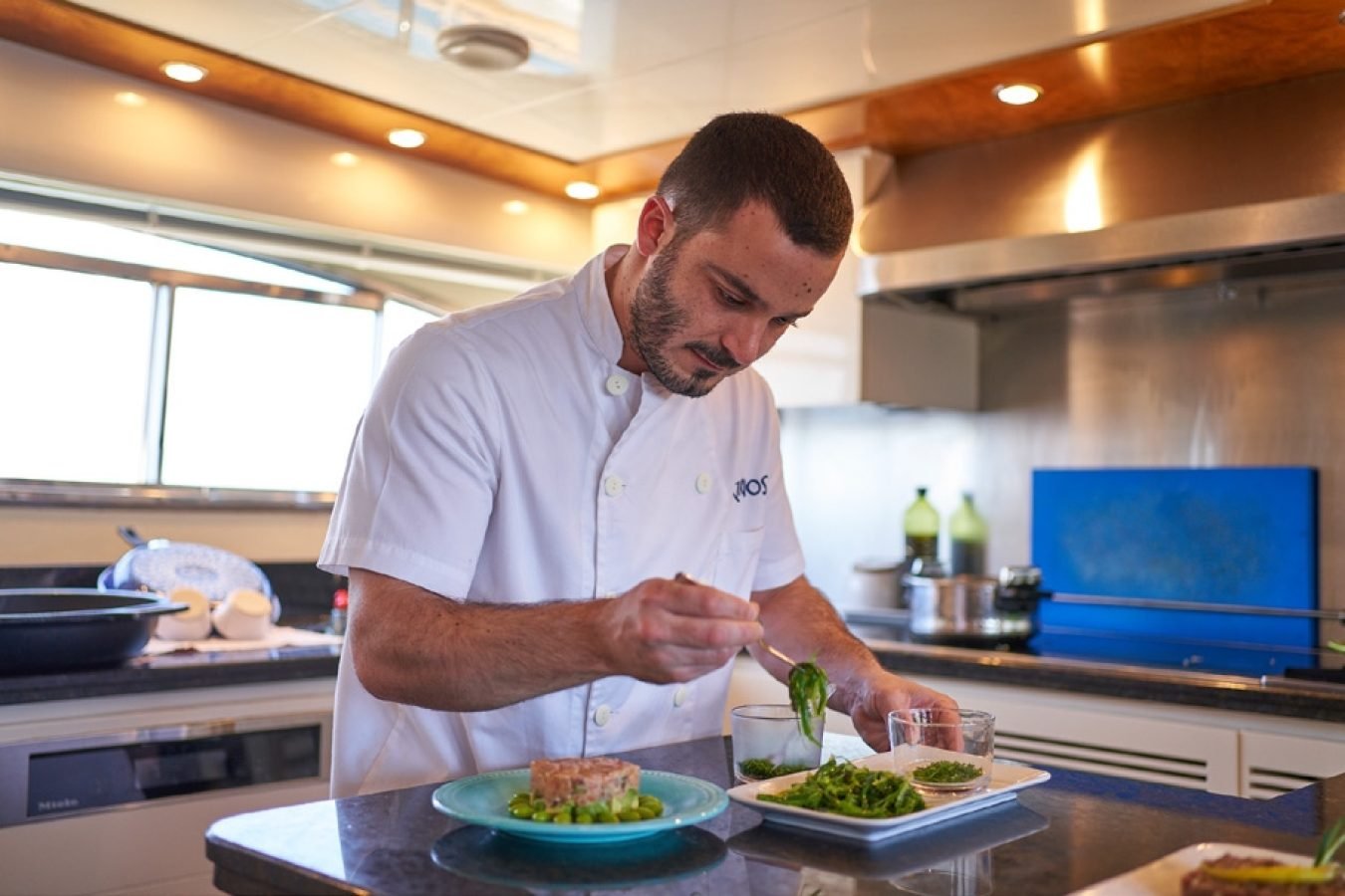 A chef in a white jacket plates food in a modern kitchen aboard the Astondoa 102 GLX My Kirios Yacht For Charter.