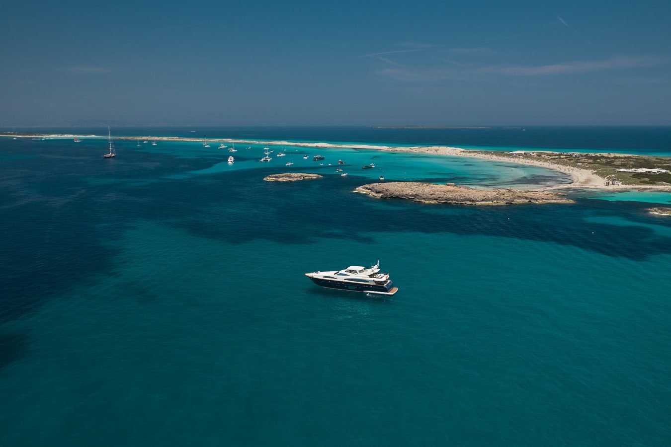 The Astondoa 102 GLX My Kirios Yacht for charter floats on clear blue water by a sandy island, boats in the distance.