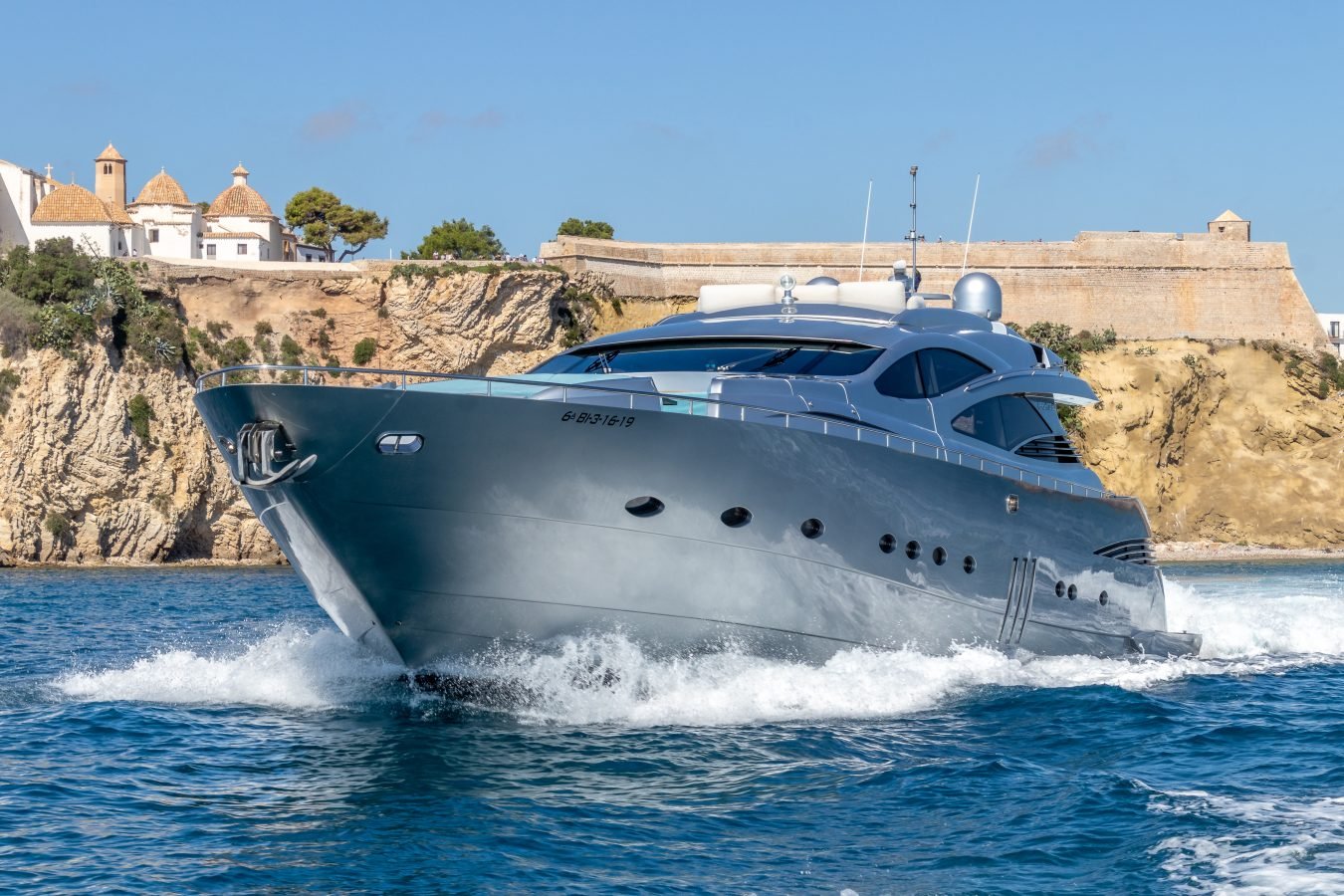 The Pershing 90 Inspiration Yacht for Charter speeds through blue water past rocky cliffs and white coastal buildings.