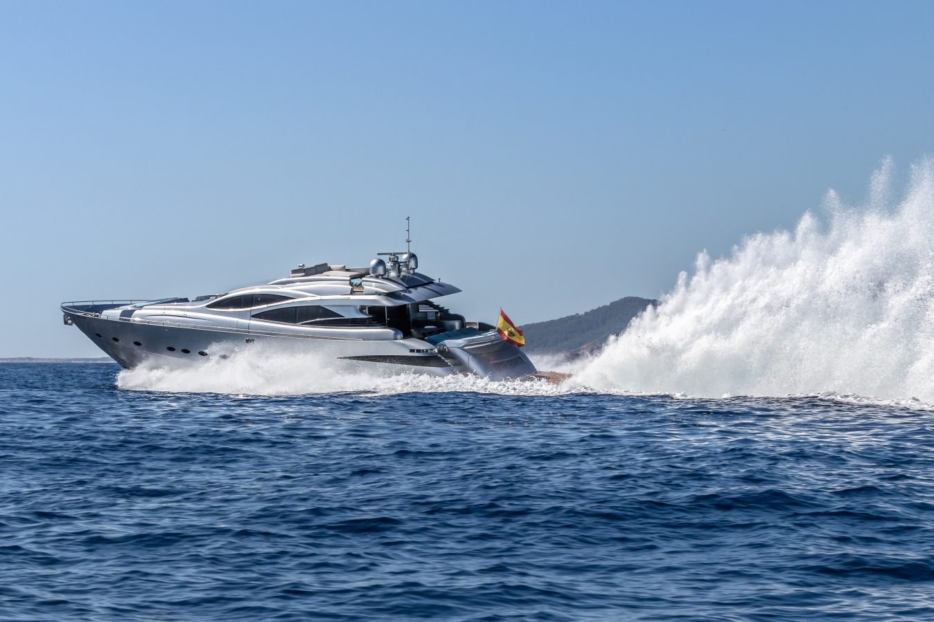 A sleek Pershing 90 Inspiration Yacht for charter speeds across blue water, splashing; hills rise in the background.