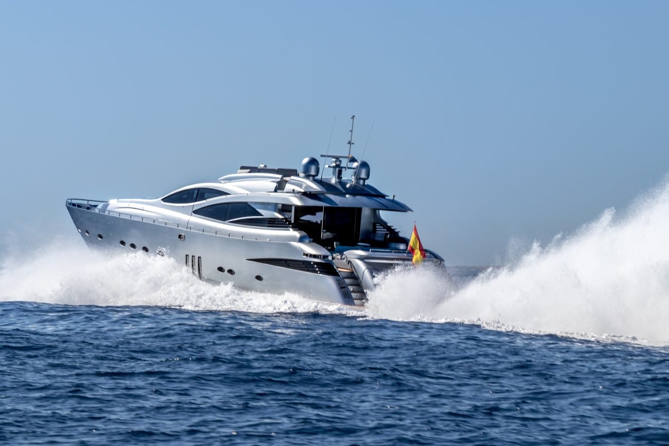 The Pershing 90 Inspiration Yacht for charter speeds over blue waves, silver hull gleaming under a clear sky and waving flag.