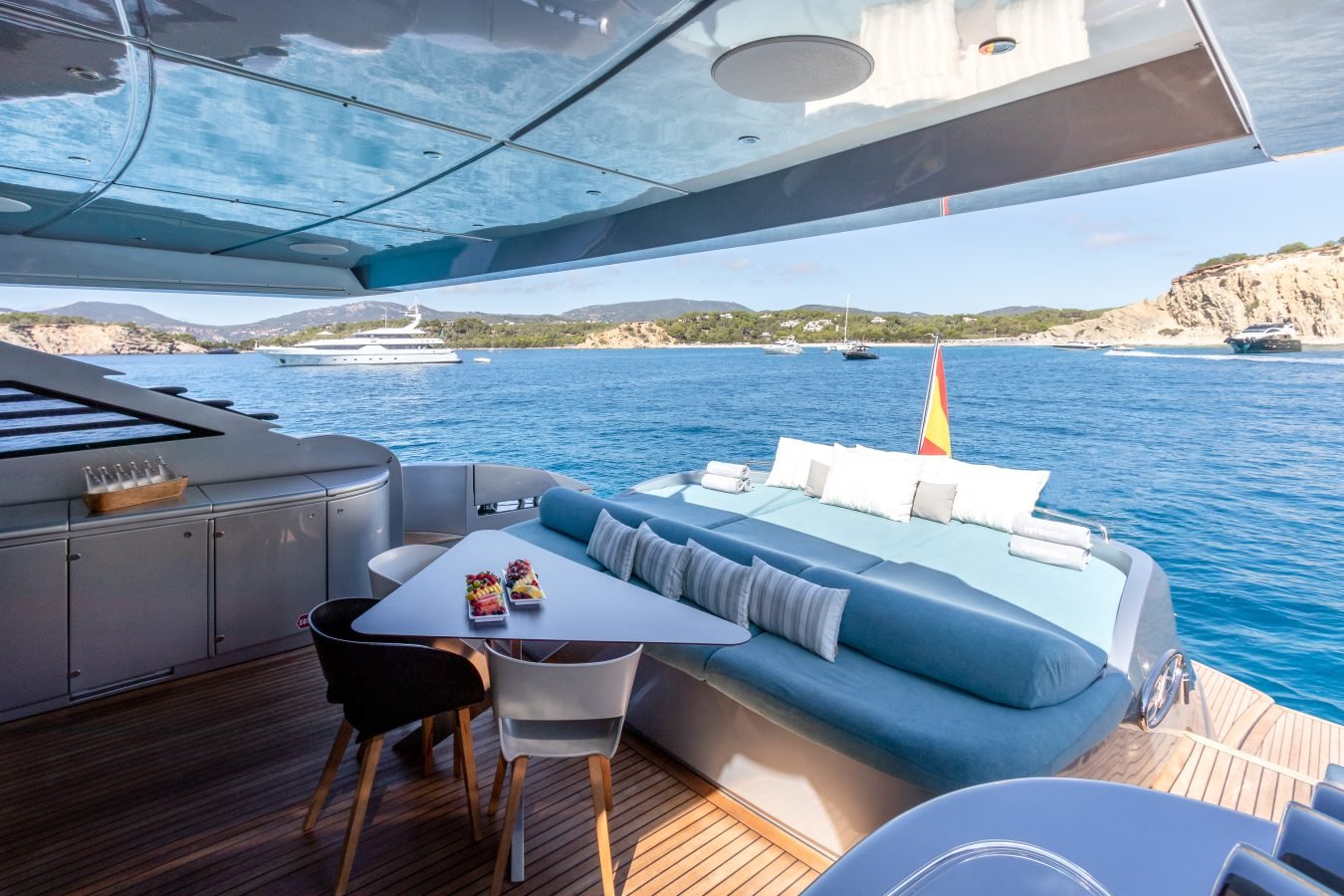 Back deck of Pershing 90 Inspiration Yacht For Charter: lounge, table with food, sea view, yachts and hills beyond.