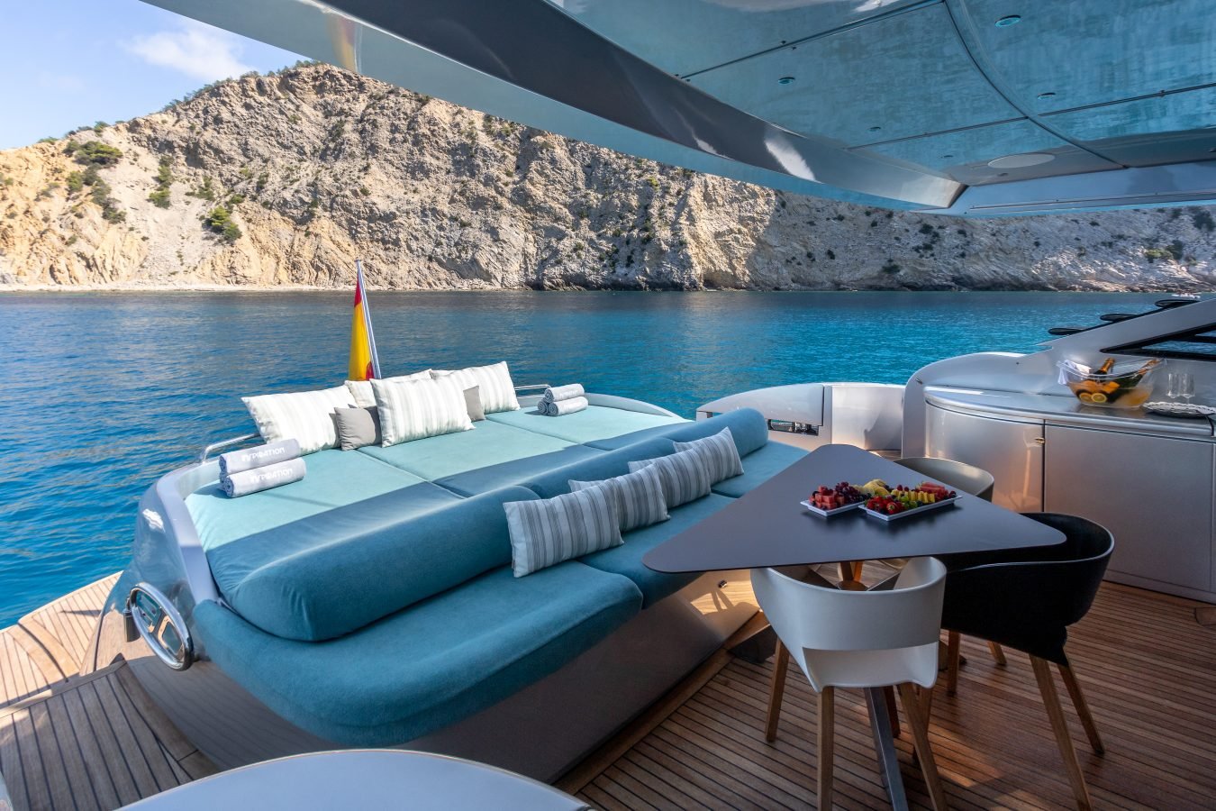 The Pershing 90 Inspiration Yacht For Charter features sun loungers, plush pillows, and a fruit platter by the coastline.