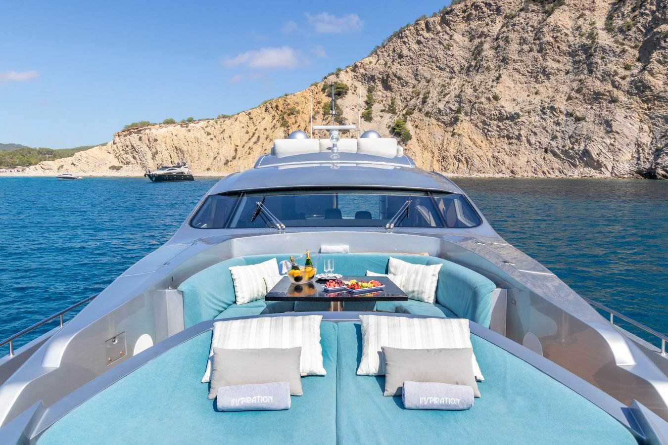 A Pershing 90 Inspiration Yacht For Charter features turquoise seating, fine dining, and stunning cliffs by blue water.