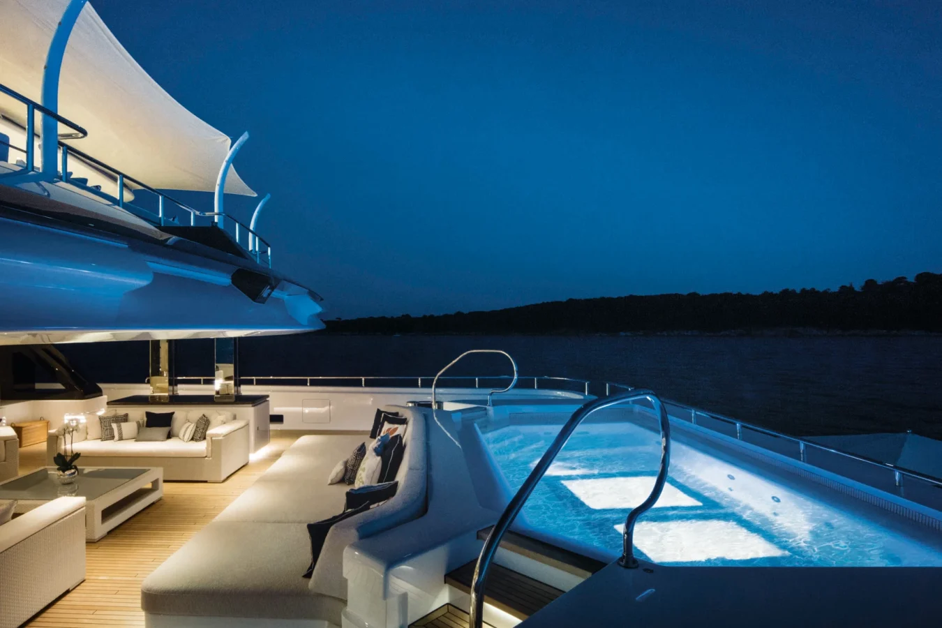 Luxury CRN Starburst IV Yacht For Charter: deck at dusk, lit pool, white sofas, modern decor, and calm sea under blue sky.