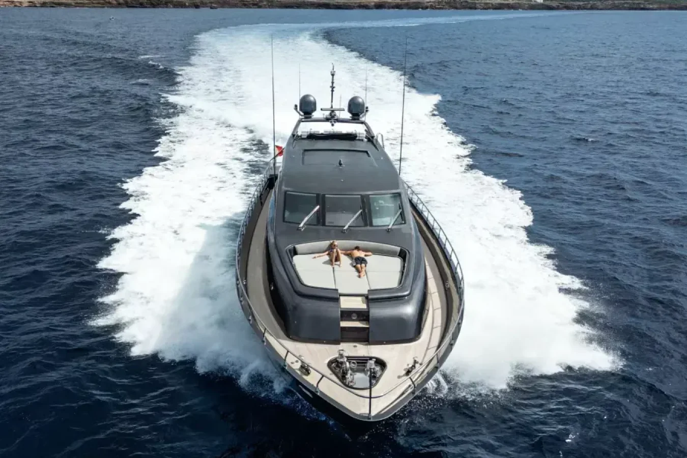 A LEOPARD 102 yacht for charter speeds through blue water as two people relax on the front deck of DARK KNIGHT Yacht.
