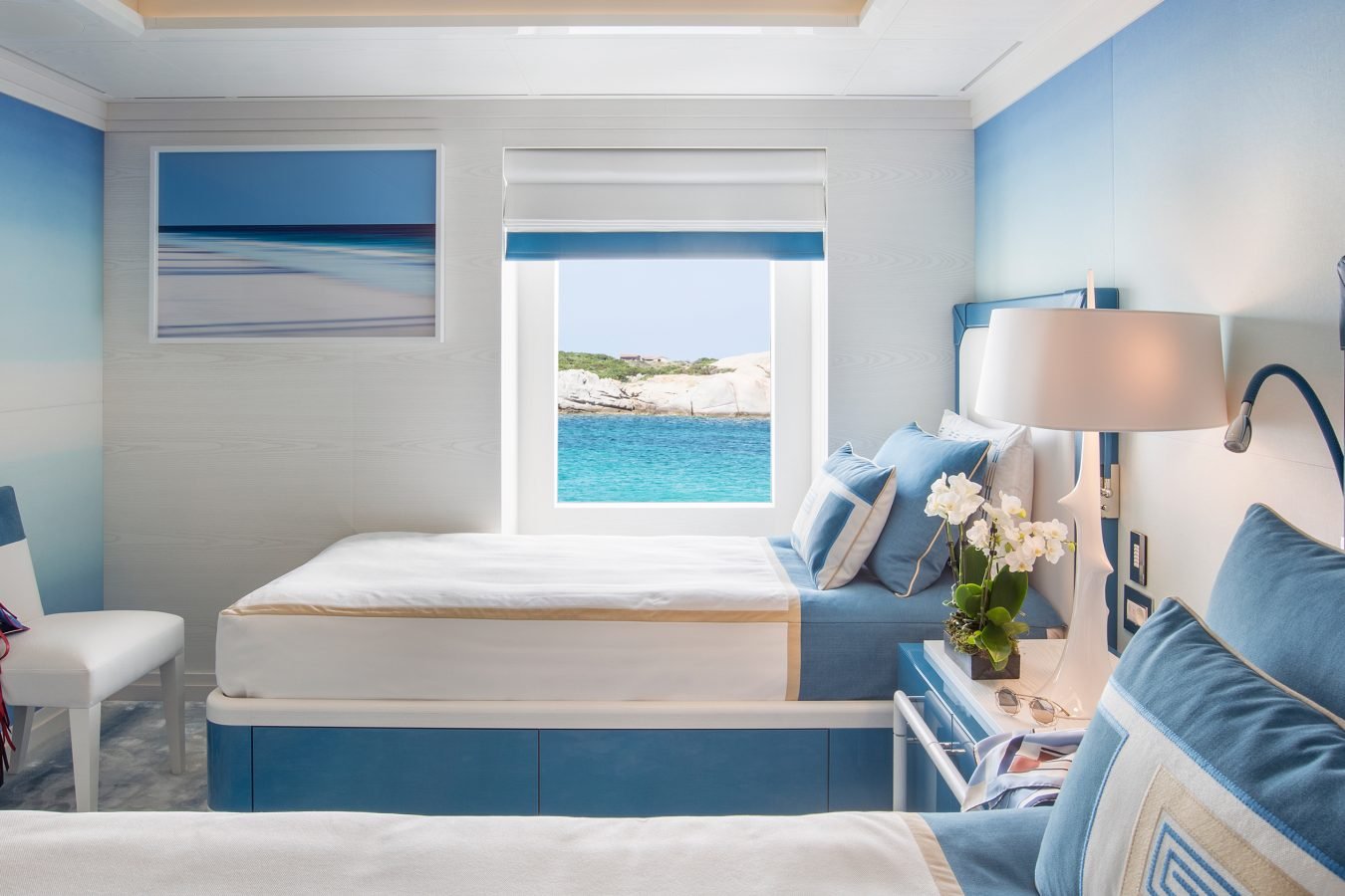 A bright, modern bedroom with twin beds, blue accents, a window showing turquoise water, and a framed beach photo.