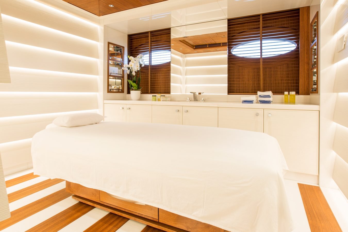 Modern spa room with a massage table, white walls, wood accents, and soft lighting; orchid on the counter.
