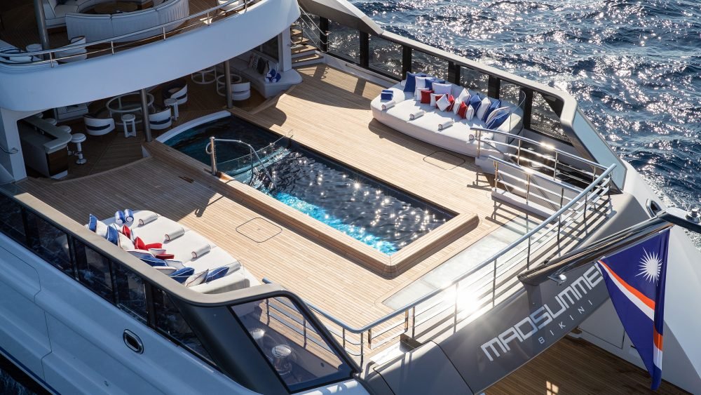 A luxury yacht deck with a pool, lounge chairs, and a flag reading MADSUMMER BIKINI above the ocean.