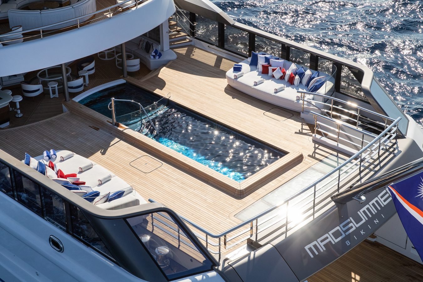 A luxury yacht deck with a pool, lounge chairs, and a flag reading MADSUMMER BIKINI above the ocean.