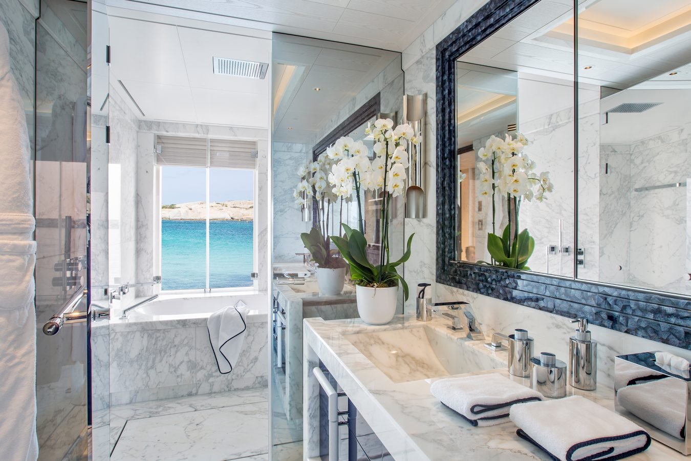 Luxurious marble bathroom with a large mirror, bathtub, orchids, and a window overlooking the ocean.