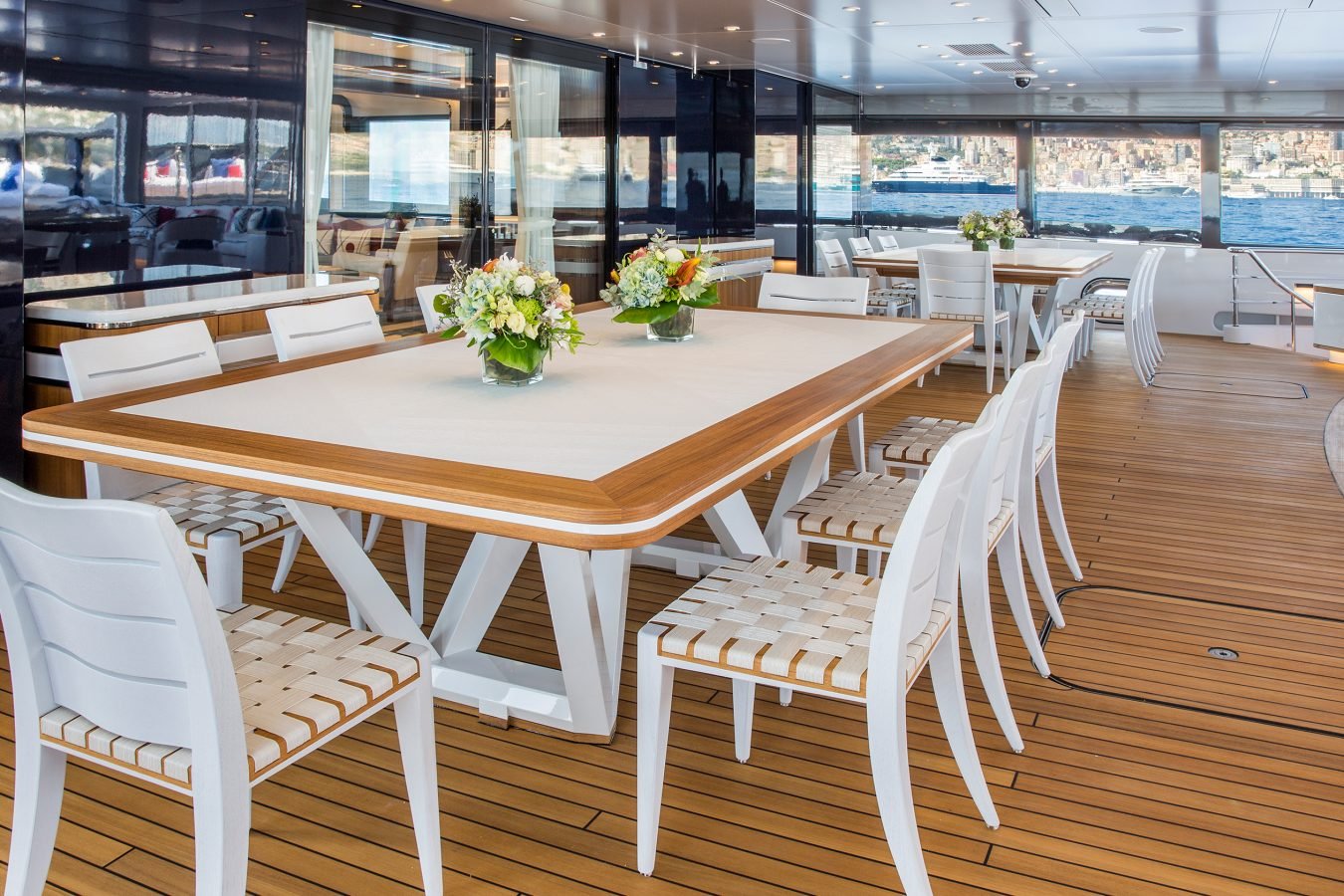Spacious yacht deck with wooden floor, white tables and chairs, floral centerpieces, and ocean view in the background.