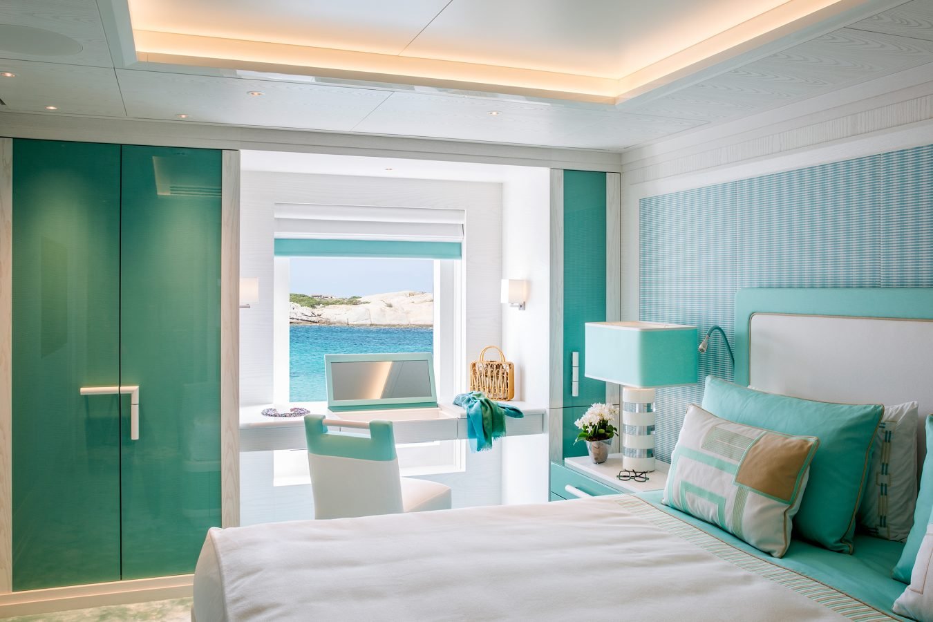 A bright, modern bedroom with teal accents, a desk by a large window, and an ocean view beyond.