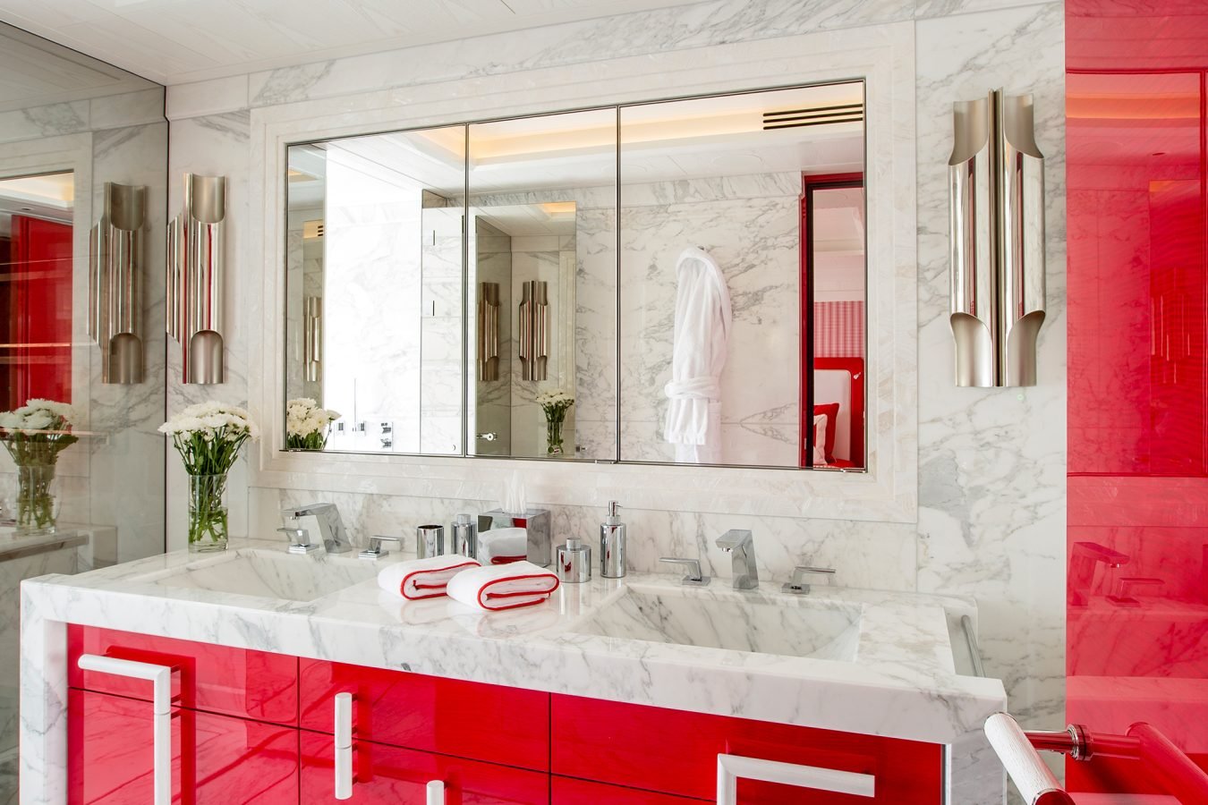 Modern bathroom with marble walls and floors, red cabinets, large mirrors, and silver light fixtures.