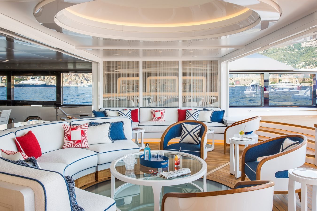Modern yacht lounge with white, blue, and red decor, curved sofas, round tables, and large windows overlooking water.