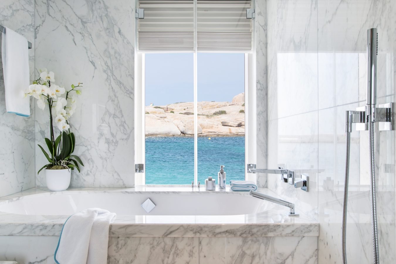 Luxurious marble bathroom with a large tub, orchid plant, window view of the sea, and modern fixtures.