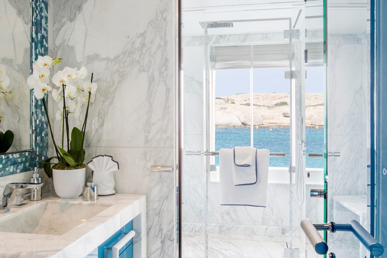 Luxurious marble bathroom with blue cabinets, glass shower, orchid plant, and a window overlooking the sea.