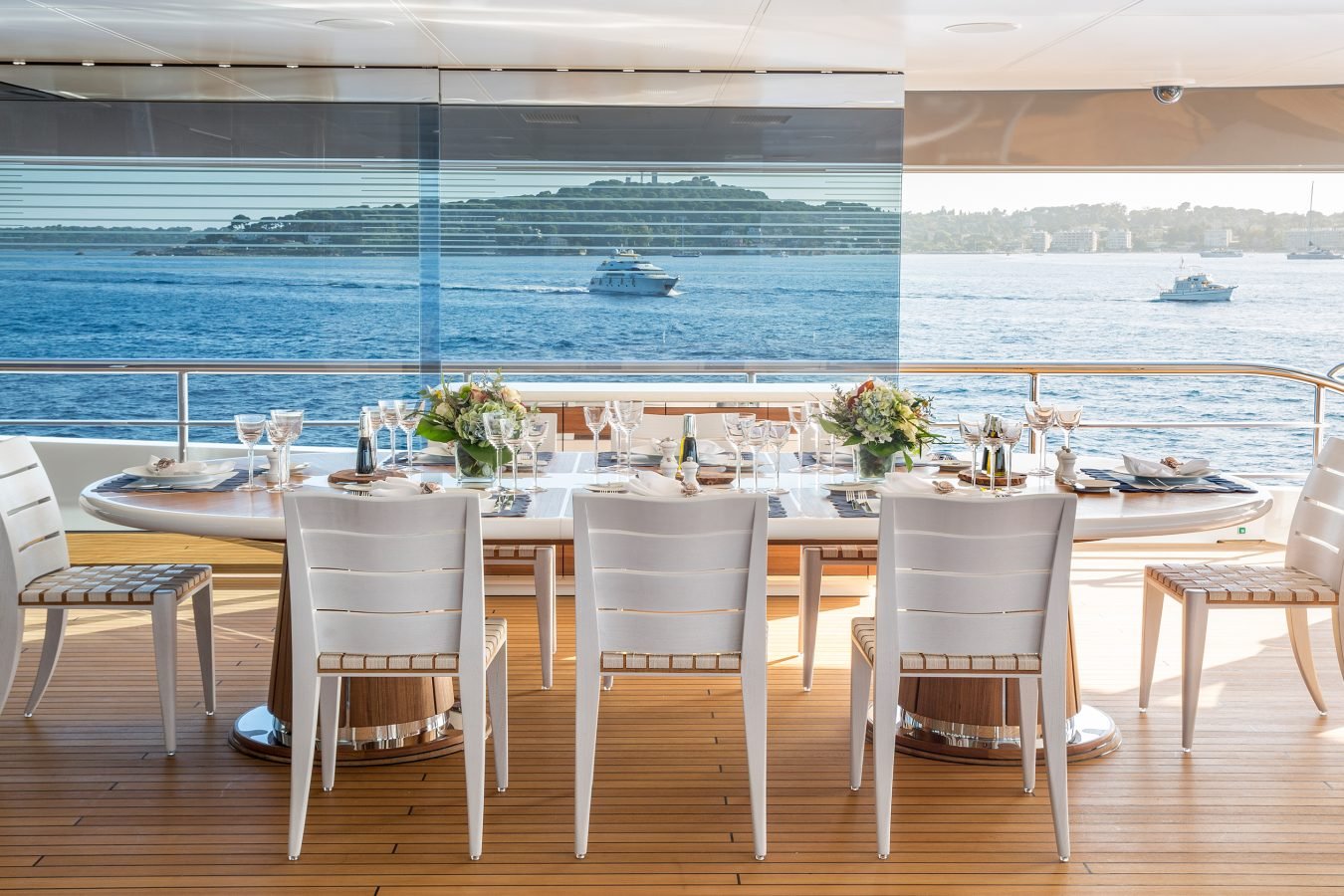 A yacht’s outdoor dining area with a set table, white chairs, and an ocean view with boats and an island in the background.