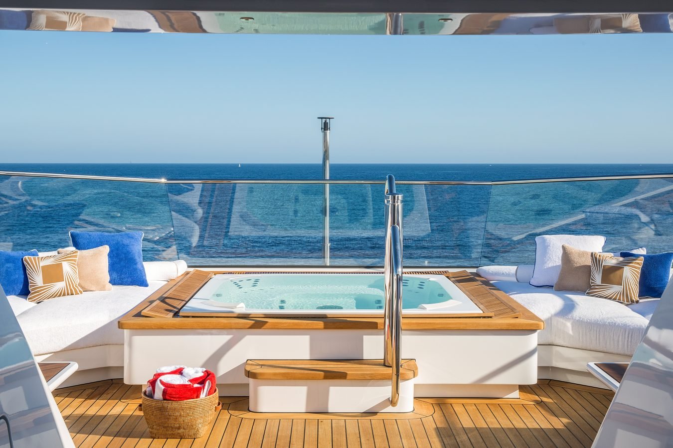 A luxury yacht deck with a hot tub, white lounge seating, blue pillows, and a view of the ocean under a clear sky.