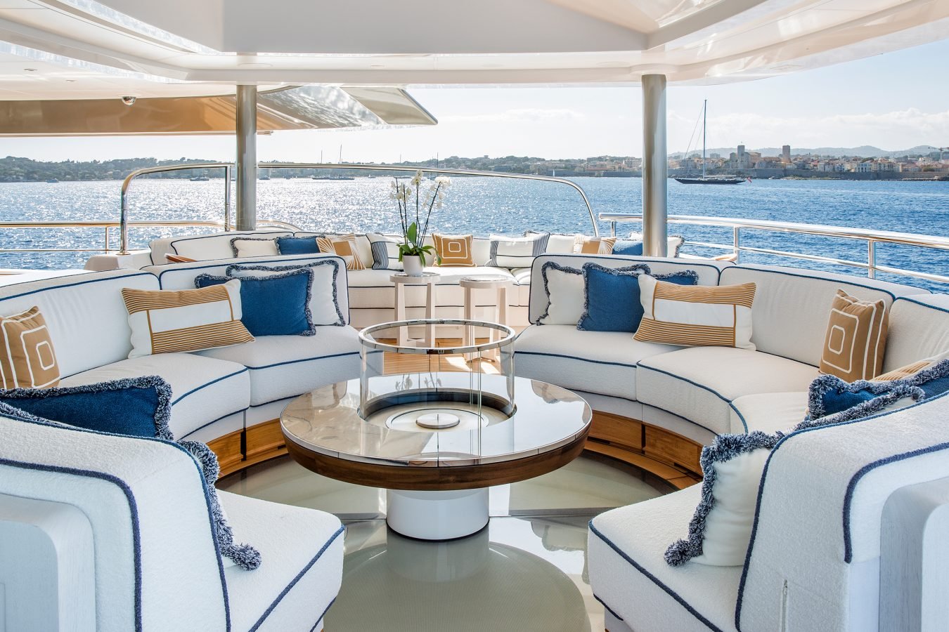 Luxurious yacht deck with curved white sofas, blue and tan pillows, glass table, and ocean view in the background.
