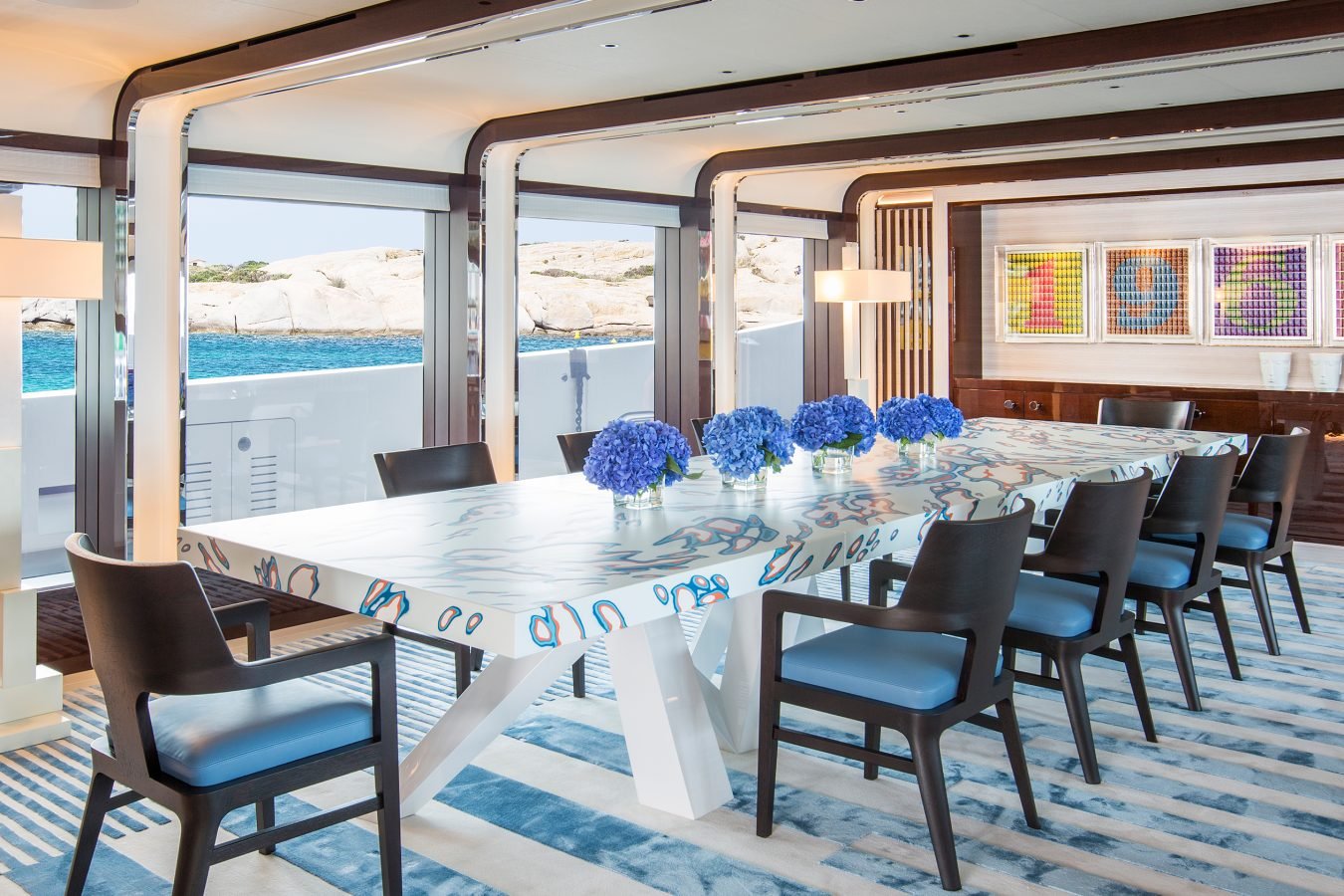 Modern dining room on a yacht with blue chairs, floral centerpieces, patterned rug, and ocean view through open windows.