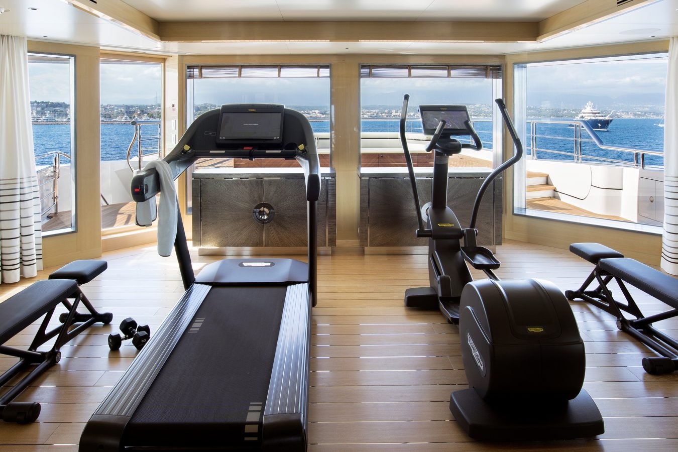 A yacht gym with treadmills, benches, and ocean views through large windows with white curtains.