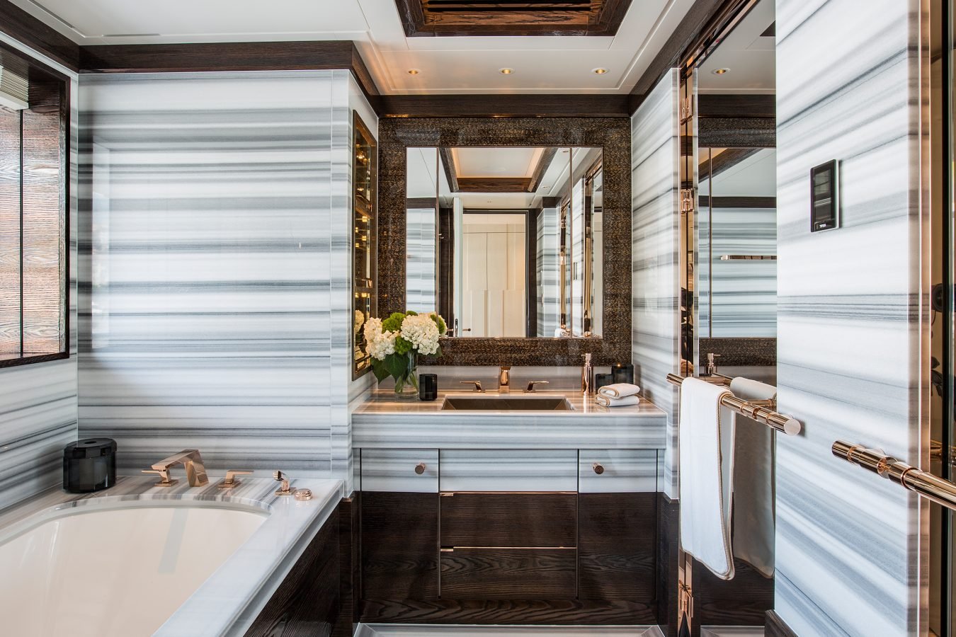 Modern bathroom with striped marble walls, large mirror, dark wood accents, bathtub, sink, and vase of white flowers.