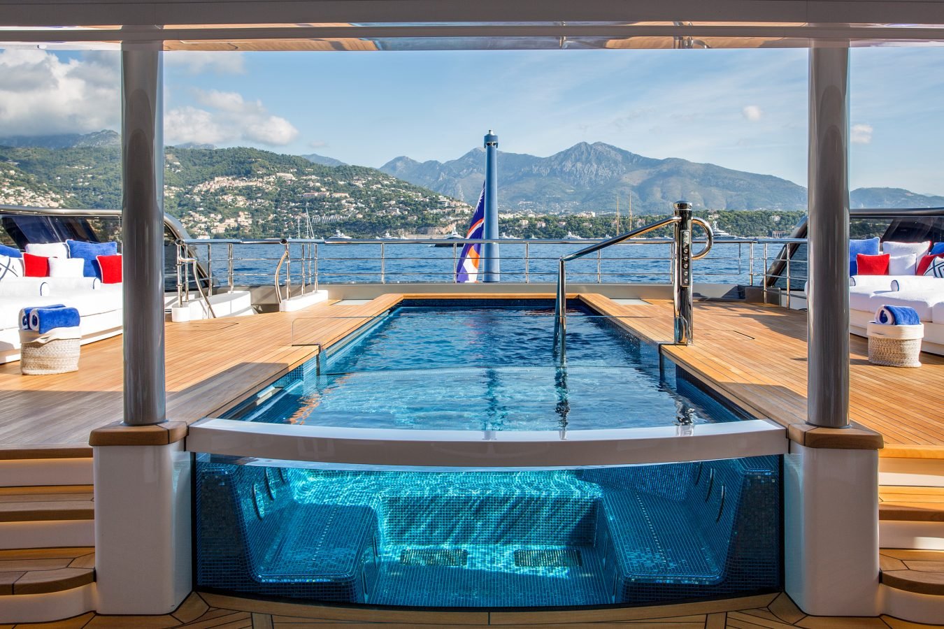 Luxury yacht deck with central glass-walled pool, lounge chairs, and scenic mountain and water views in the background.