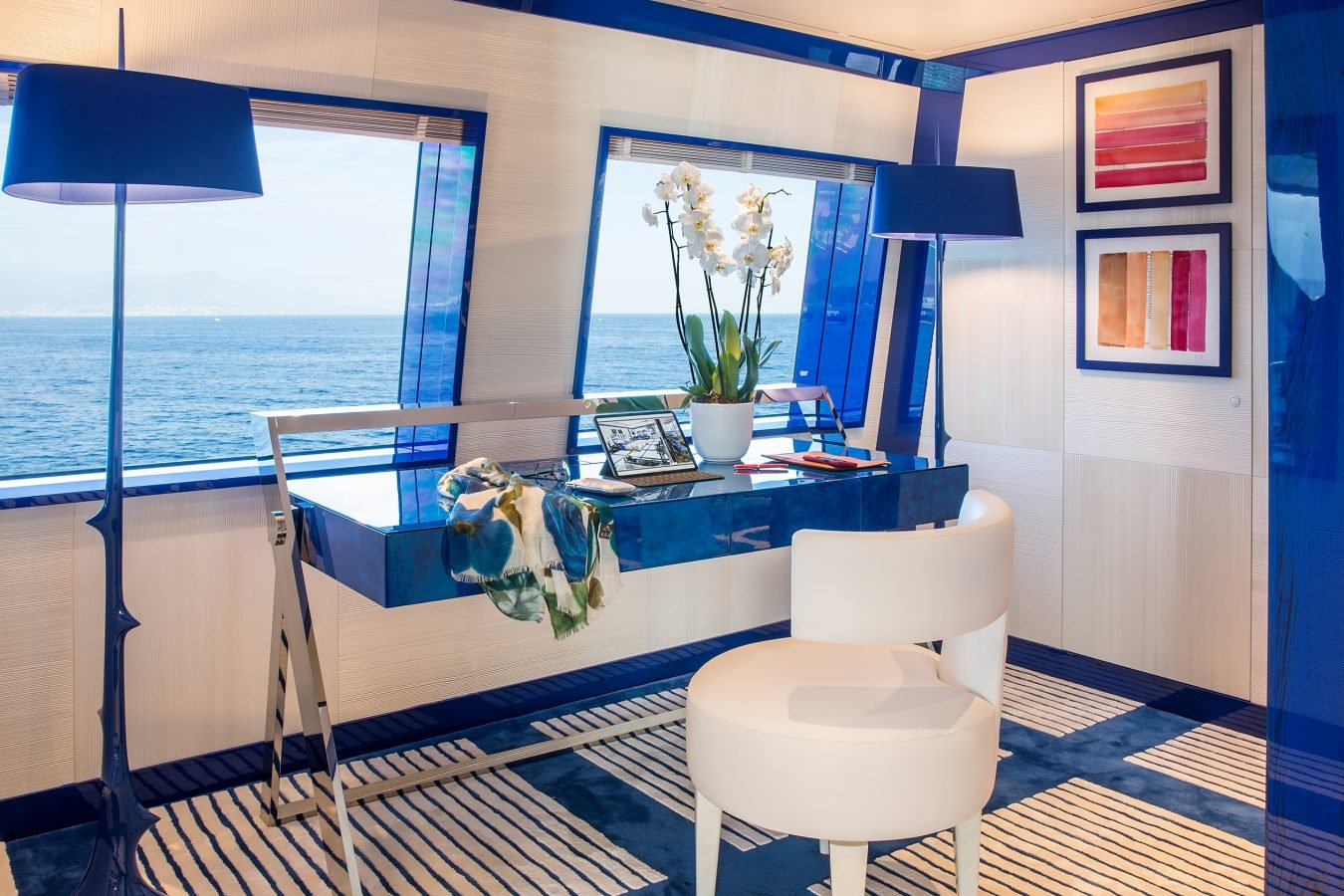 Modern office with ocean view, blue accents, desk with laptop, white chair, orchids, and framed art on the wall.