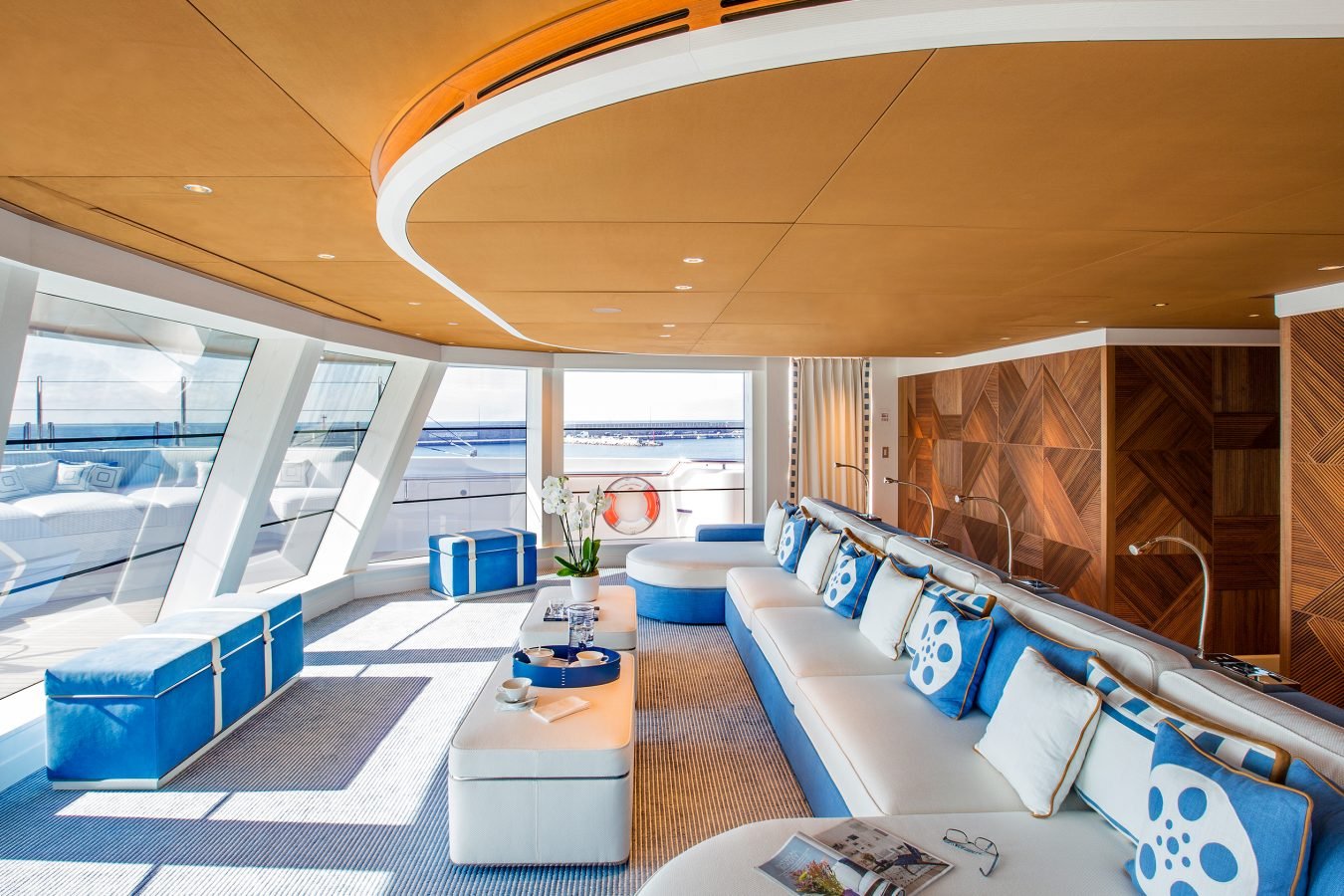 Modern yacht lounge with blue and white seating, geometric pillows, large windows, and wooden ceiling and walls.