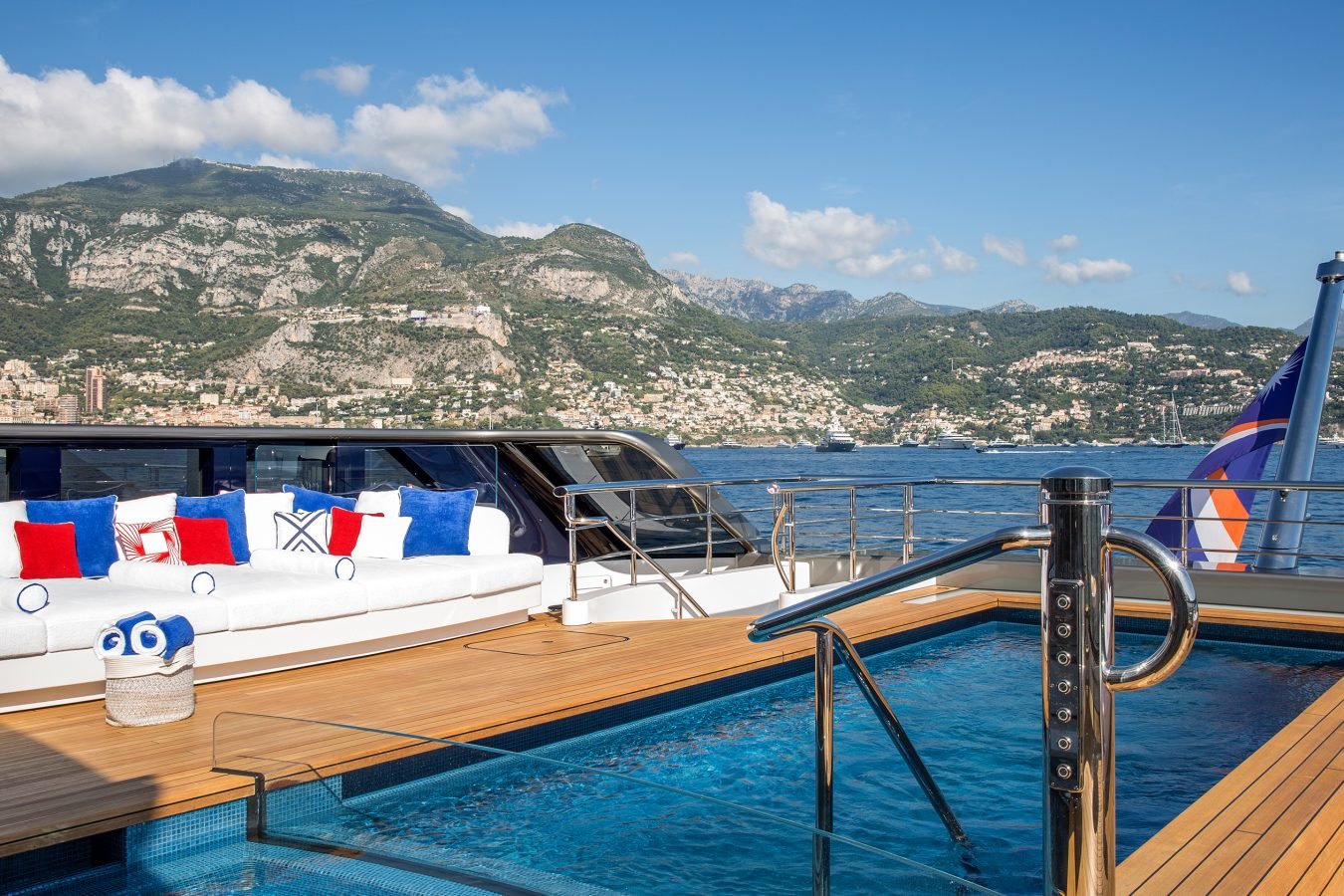 Luxurious yacht deck with pool, white lounge seating, cushions, basket, sea, and mountains in the background.