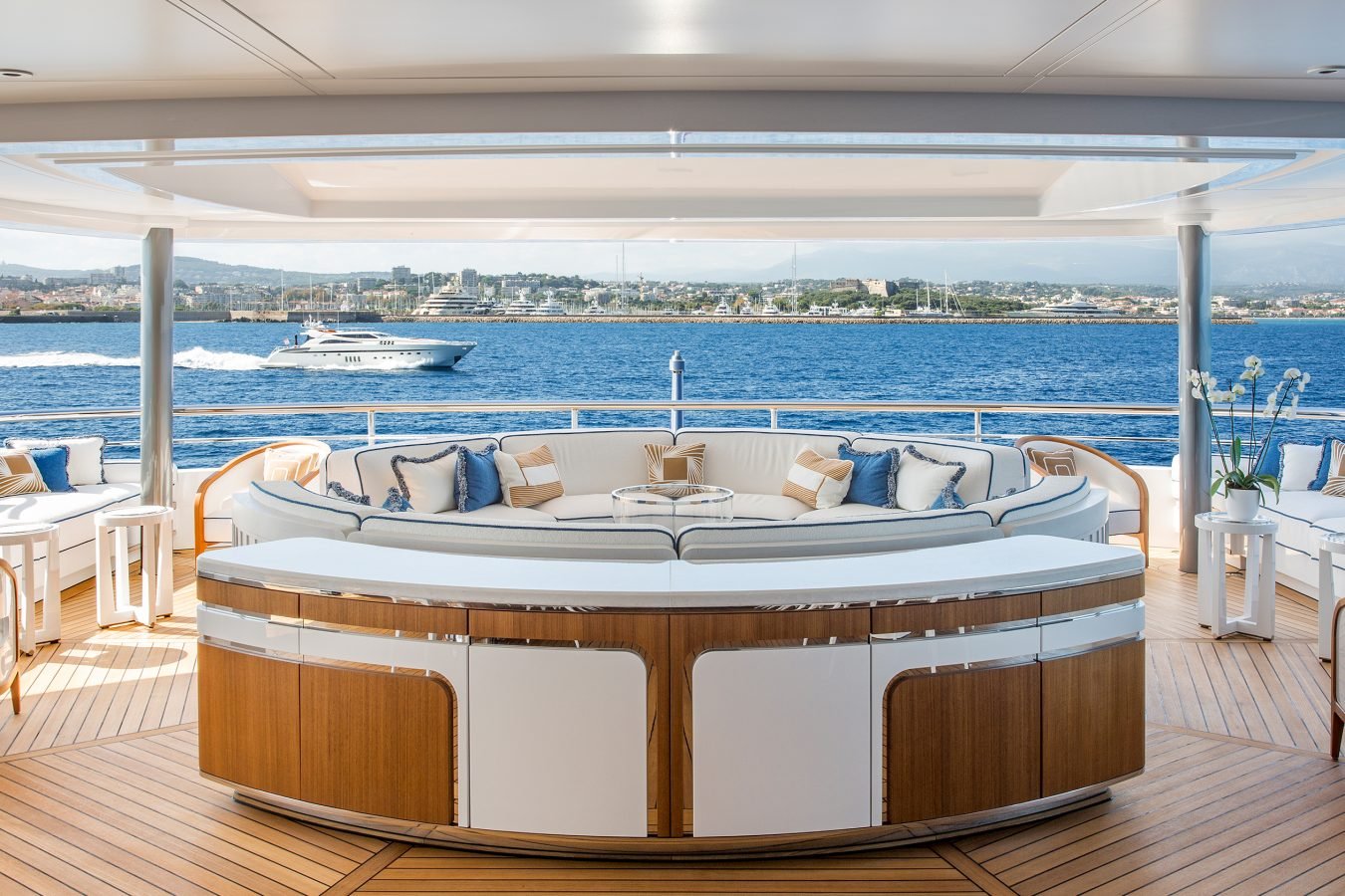 Luxurious yacht deck with curved sofa, ocean view, city skyline, and boat speeding by in the background.